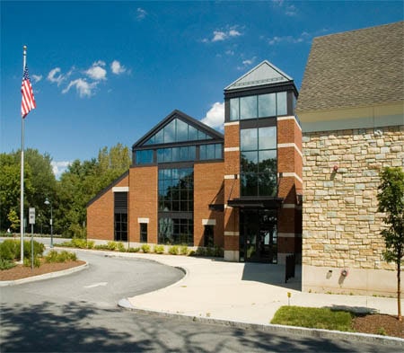 A modern brick and glass building with large windows, an American flag on a pole in front, surrounded by trees and a clear blue sky.