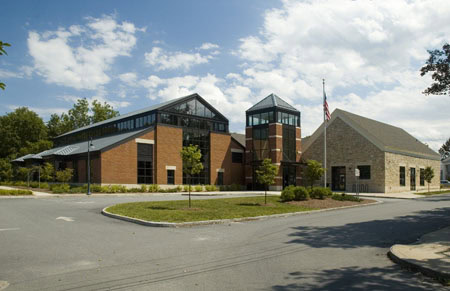A modern building with red brick and stone walls, large windows, and a sloped metal roof, surrounded by trees and greenery under a partly cloudy sky. An American flag is visible near the entrance.
