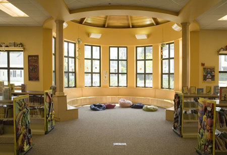 A brightly lit children’s reading area in a library with tall windows, colorful bean bags on the floor, and bookshelves decorated with illustrated panels.