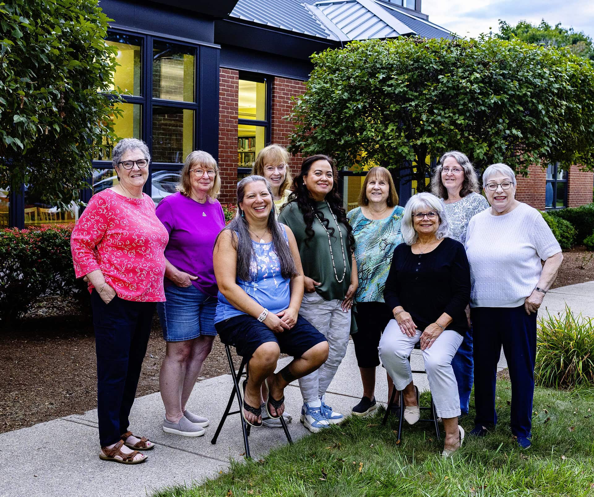 Nine women pose and smile together outside a building, surrounded by green bushes and trees. Some are standing, and some are sitting on chairs, with a pathway and windows visible in the background.